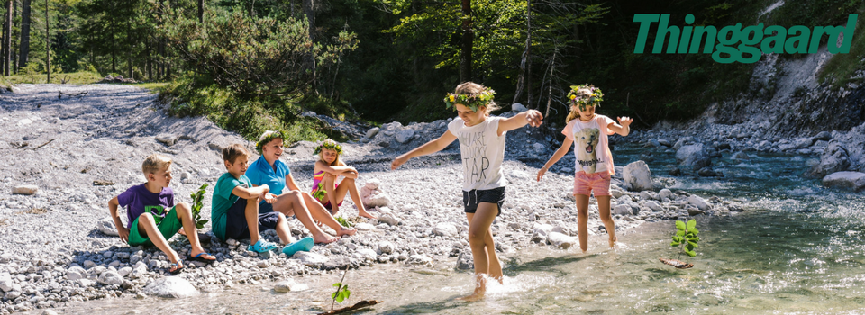 Børn leger ved en stenet å på en solrig sommerferie Østrigsdag. Nogle sidder på bredden, mens to går gennem det lave vand. De bærer blomstrede pandebånd og smiler glad. Træer og grønne områder omgiver området. Ordet Thinggaard er synligt i øverste højre hjørne.