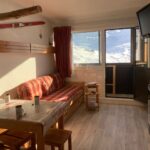 En hyggelig stue med en rød plaid sofa, et træbord og to stole. En ski fra Westendorf pynter på væggen. Sollys strømmer gennem et vindue og afslører en sneklædt udsigt over bjergene. Et fladskærms-tv er monteret på væggen.