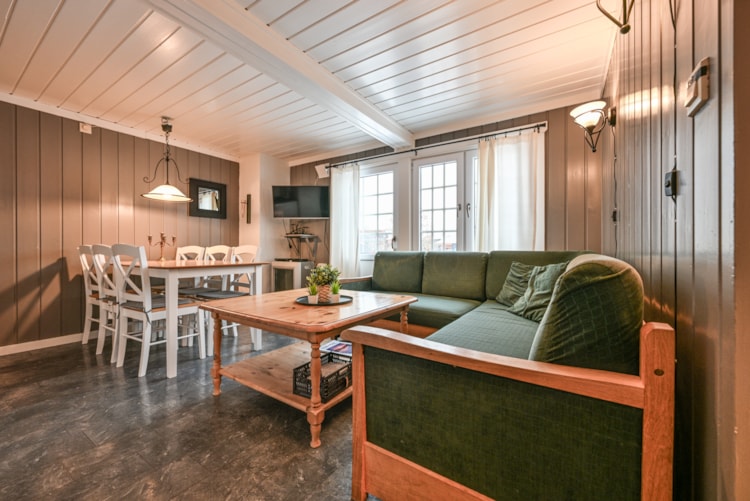 Hyggelig stue med en grøn L-formet sofa, sofabord i træ og et spisebord med stole. Værelset har træpanelvægge og bjælker, hvilket fremkalder en skiferie-stemning. Store vinduer med gardiner giver udsigt, der minder om Wagrain. Et lille TV er monteret på væggen.