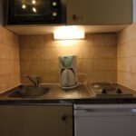Et kompakt tekøkken i Wagrain har en metalvask med vandhane, et elektrisk komfur med to brændere og et lille hvidt køleskab. En sort mikroovn står på en hylde. Under kabinettet er en hvid kaffemaskine, oplyst af lys, med solbrune fliser, der danner bagpladen - perfekt til din skiferie-flugt.