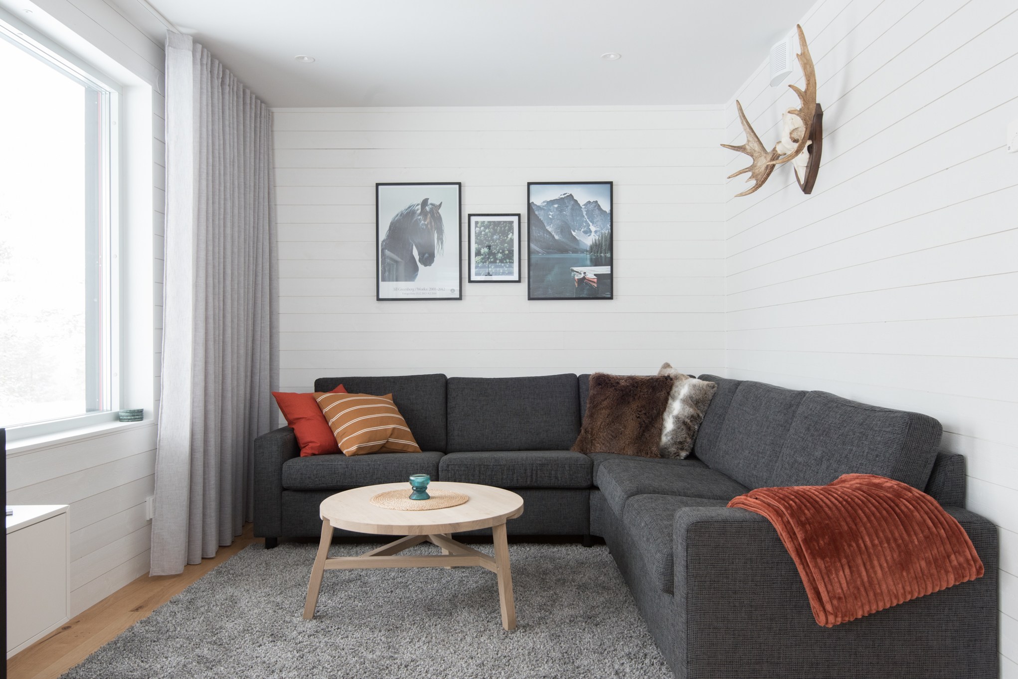 En moderne stue med en mørkegrå sektionssofa prydet med orange og brune puder fremkalder Westendorfs hyggelige charme. Et rundt sofabord i træ står på et gråt tæppe, mens tre indrammede billeder og gevirer pryder de hvide træpaneler, der minder om skirejseeventyr.