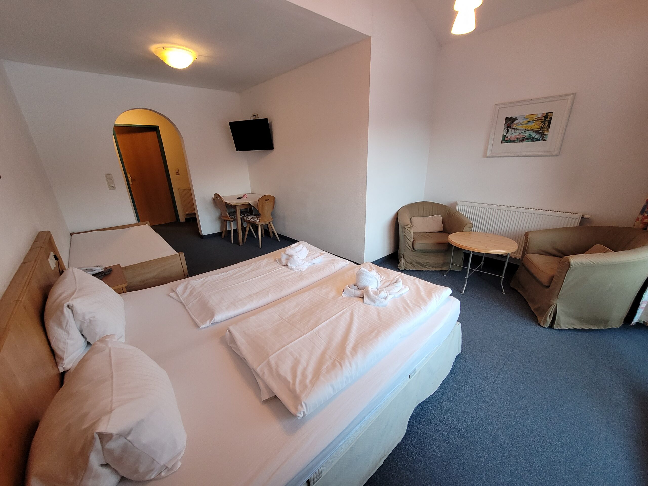 Et hotelværelse med en pænt redt dobbeltseng, et lille bord med to stole, et fladskærms-tv monteret på væggen og en ekstra enkeltseng i hjørnet. Værelset har hvide vægge og gråt tæppe.
