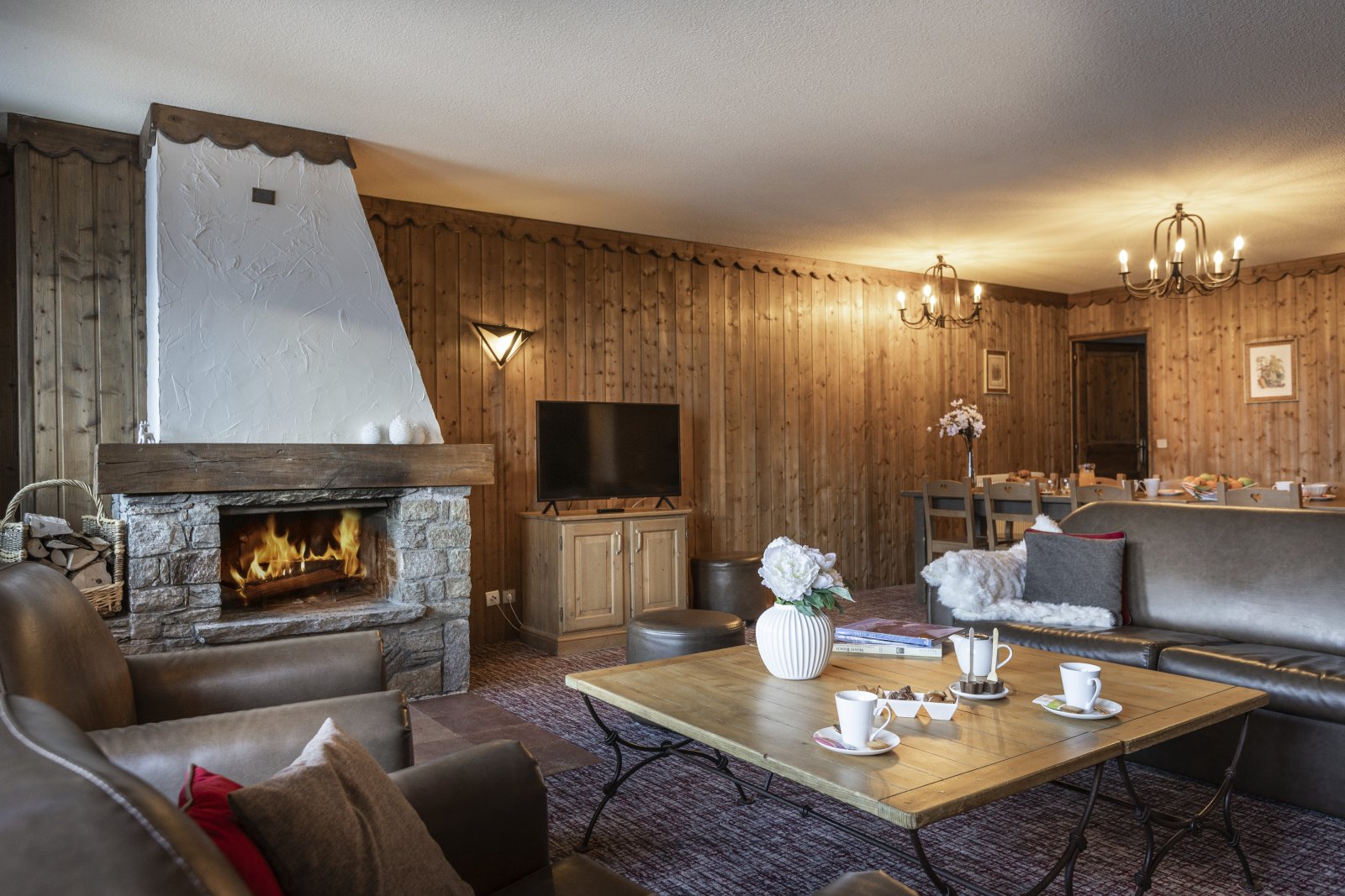 Hyggelig hytteindretning med trævægge og loft i naturskønne Saalbach. En stenpejs er tændt ved siden af et fladskærms-tv. Lædersofaer og et træsofabord med tekopper står i forgrunden, mens et spisebord venter under lysekronebelysning - et perfekt tilflugtssted for skirejser.