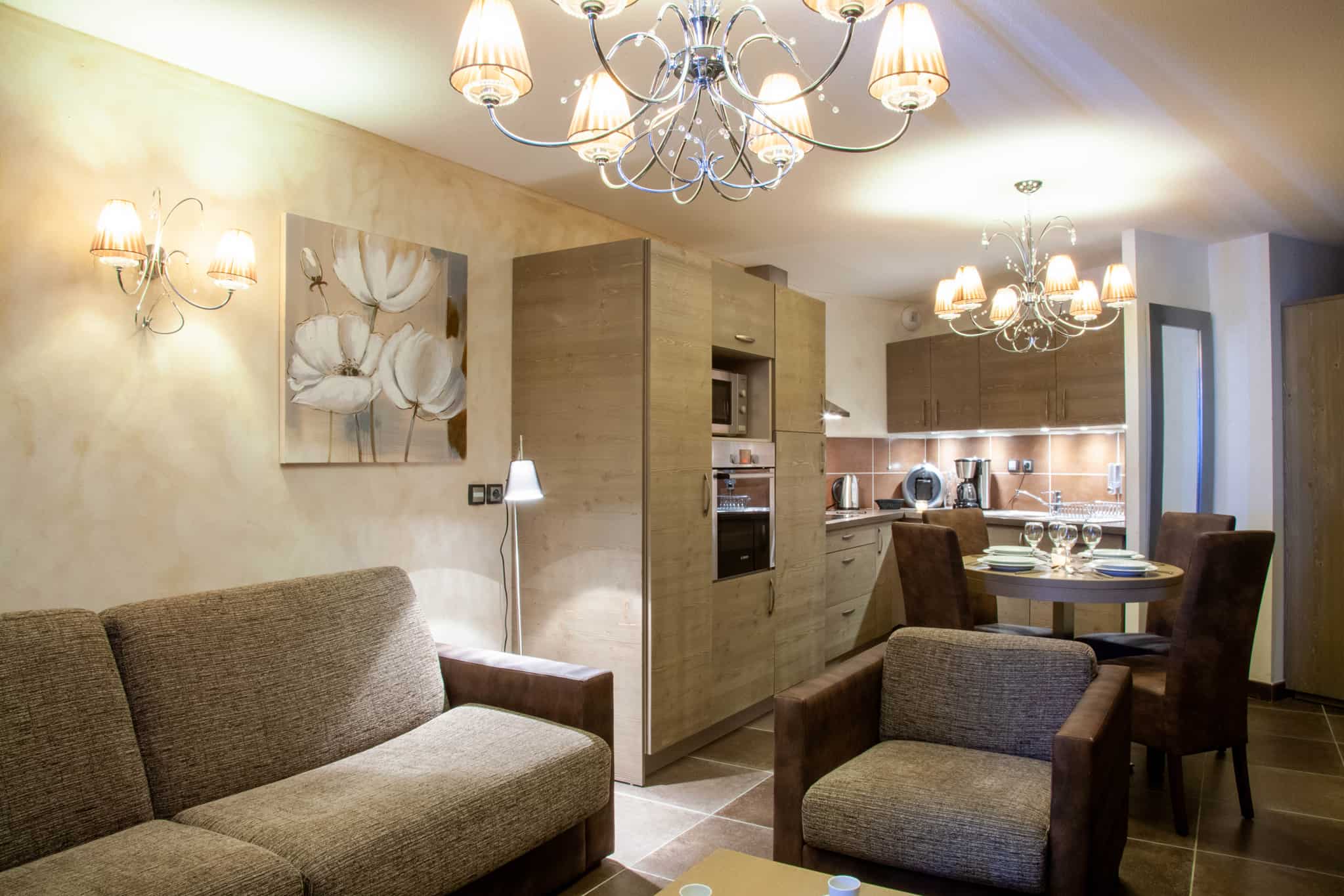 En moderne stue og køkkenområde med beige og brune toner fremkalder et strejf af Westendorf-charme. Rummet byder på sofa, lænestole, rundt spisebord med vinglas, træskabe og elegante lysekroner. Et blomstermaleri pryder væggen nær det hyggelige opholdsområde.