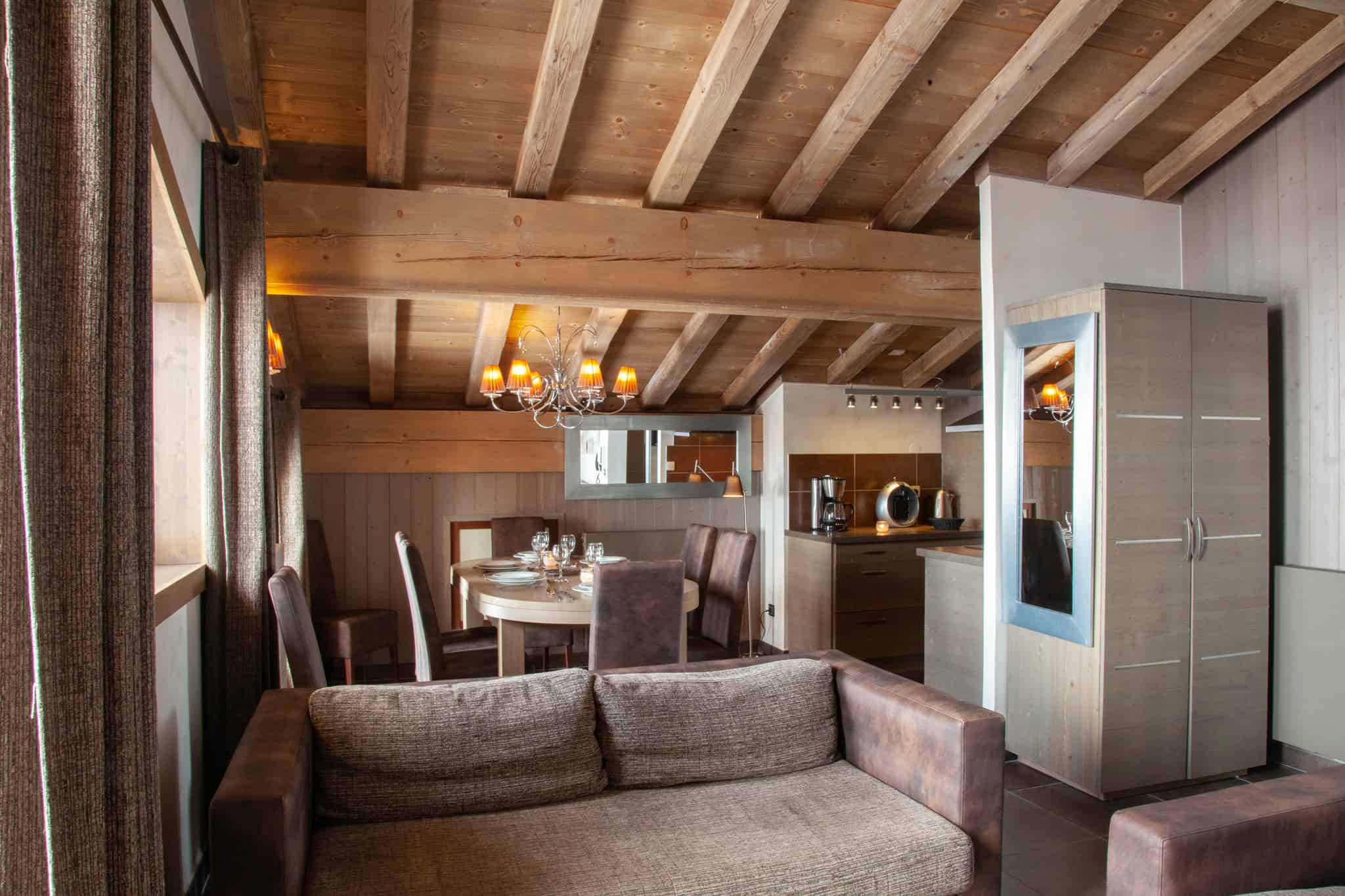 Hyggeligt opholdsrum i hjertet af Wagrain, med synlige træbjælker, en brun sofa og et spisebord med stole ved siden af et moderne tekøkken. Varm belysning fra en lysekrone skaber en indbydende atmosfære, perfekt til afterski-afslapning, når du nyder nærliggende skirejsers eventyr.