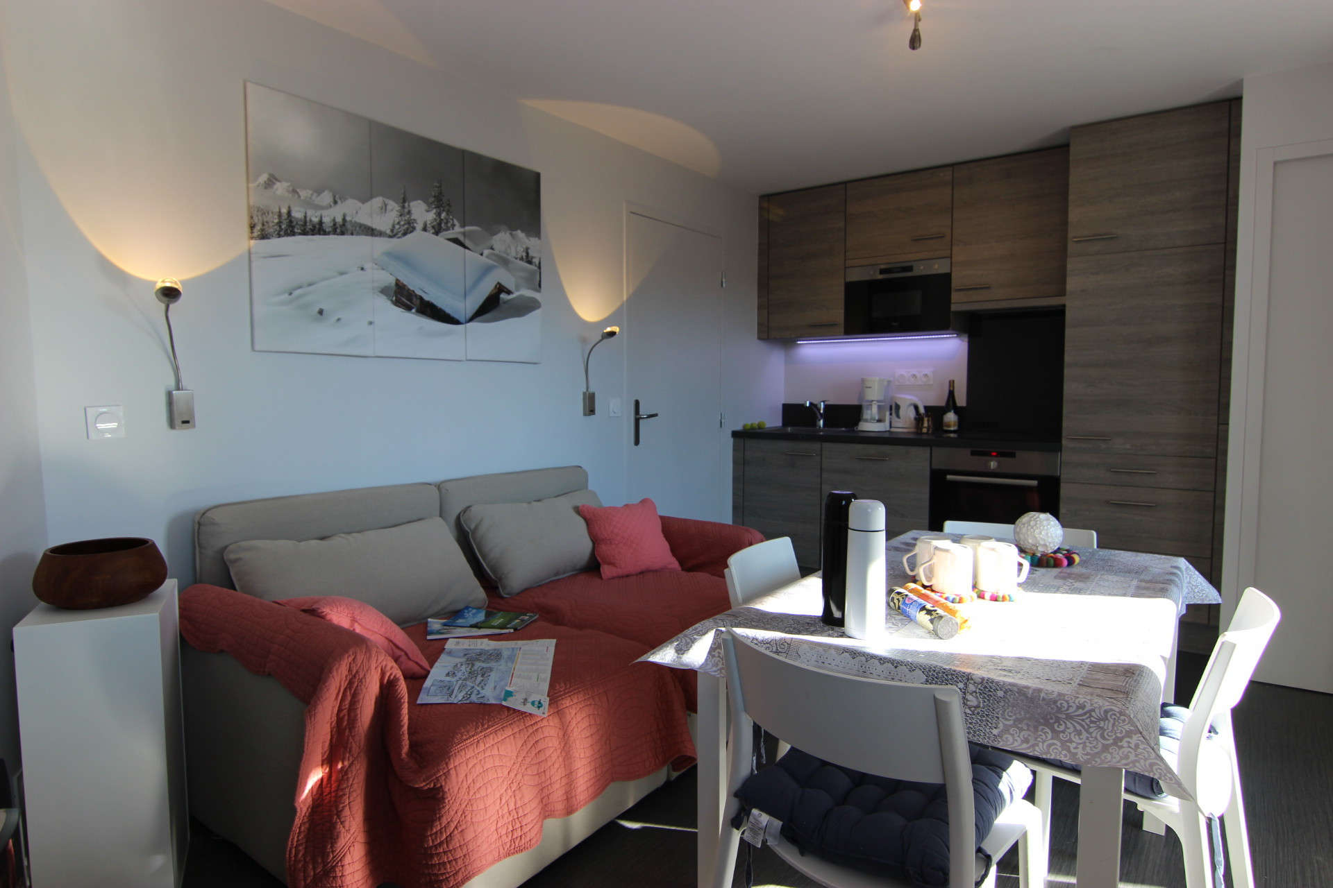 En hyggelig stue med en grå sofa beklædt med et rødt træk, et bord dækket med en mønstret klud og køkkenskabe i baggrunden. Et landskabsfoto af sneklædte Saalbach-bjerge hænger på væggen, og to væglamper oplyser rummet.