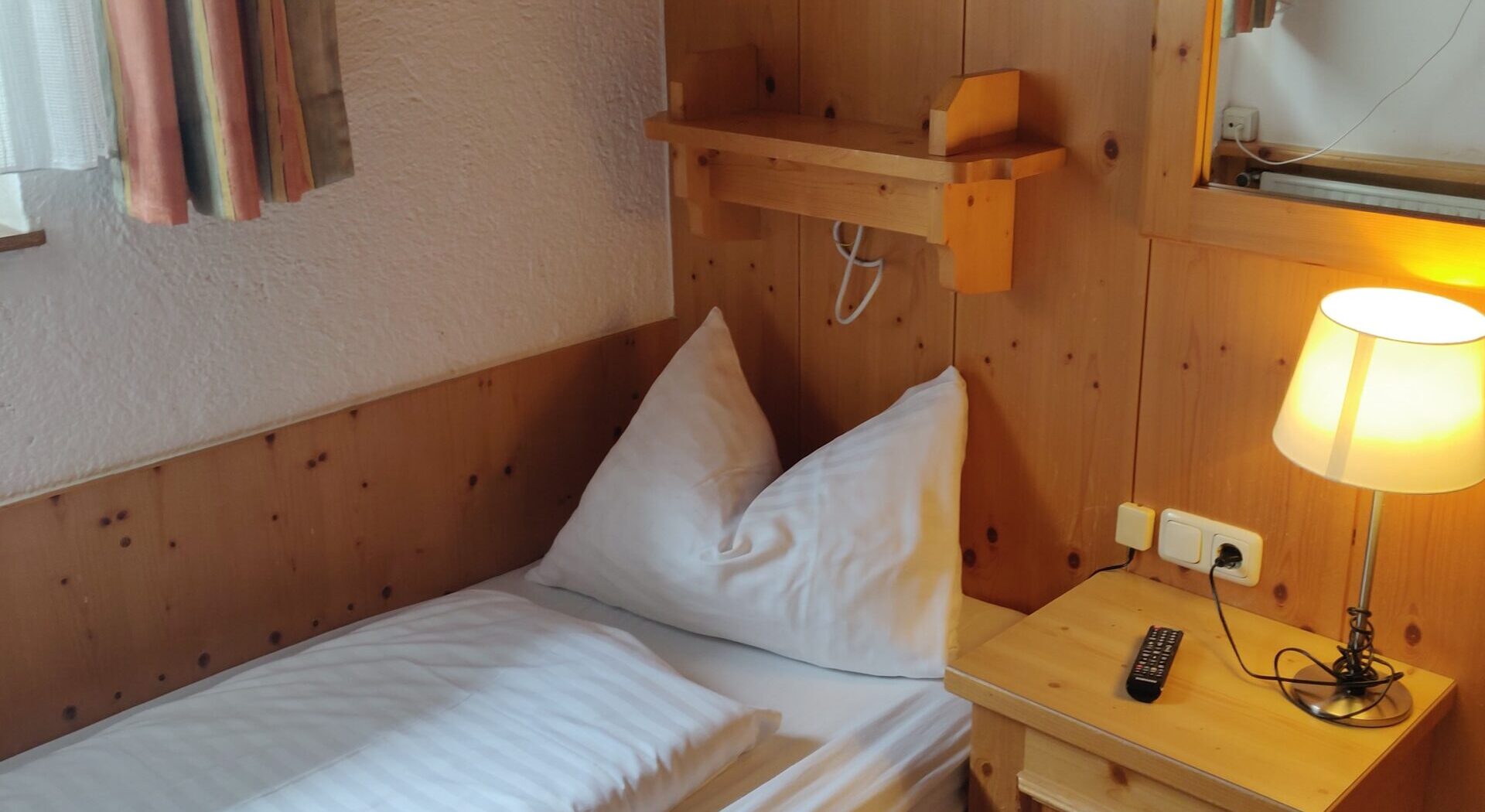 En hyggelig enkeltseng med hvidt sengetøj ved siden af et trænatbord med en lampe og fjernbetjening, ved siden af et vindue med gardiner. Et spejl og et bjergmaleri hænger på træpanelvægge.
