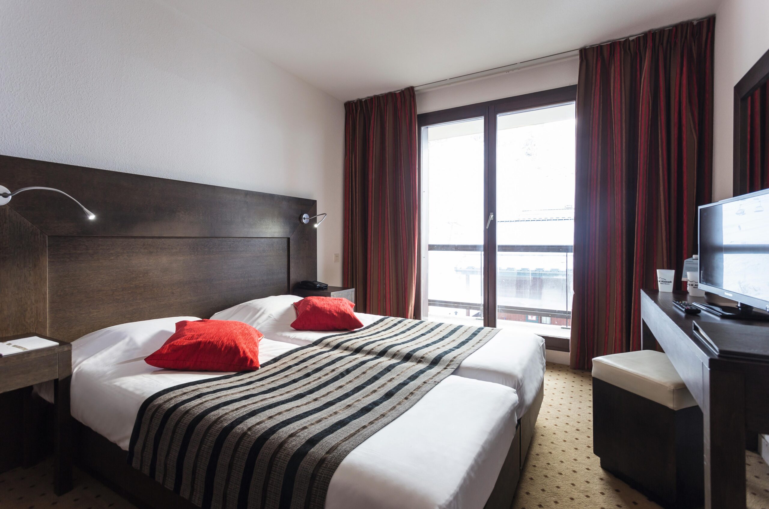 Hotelværelset, perfekt til en skiferie i Saalbach, har to pænt arrangerede enkeltsenge med hvidt sengetøj, sortstribede løbere og røde puder. Et stort vindue med røde gardiner lukker naturligt lys ind. Et tv står på et skrivebord i mørkt træ ved siden af en stol på et mønstret tæppe.