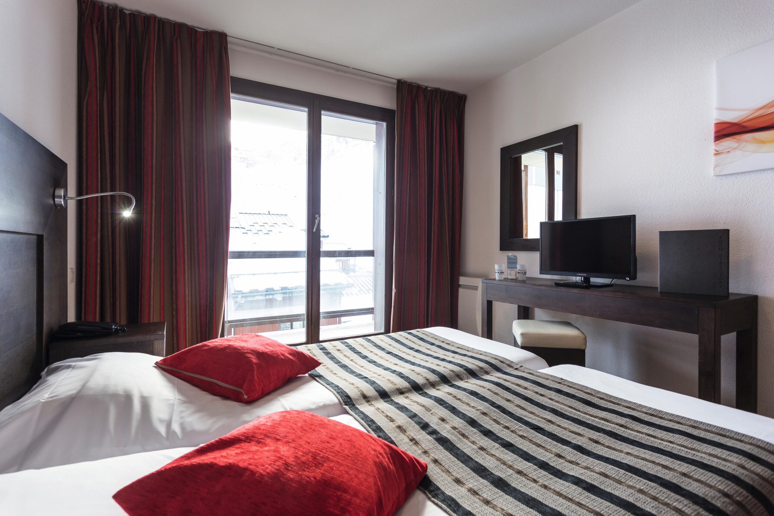 Hotelværelse med to senge, hver prydet med en rød pude og stribet løber. Et træskrivebord med tv og spejl er til højre. Et stort vindue med røde gardiner lukker naturligt lys ind og afslører en balkonudsigt, der minder om en sommerflugt til Saalbach.