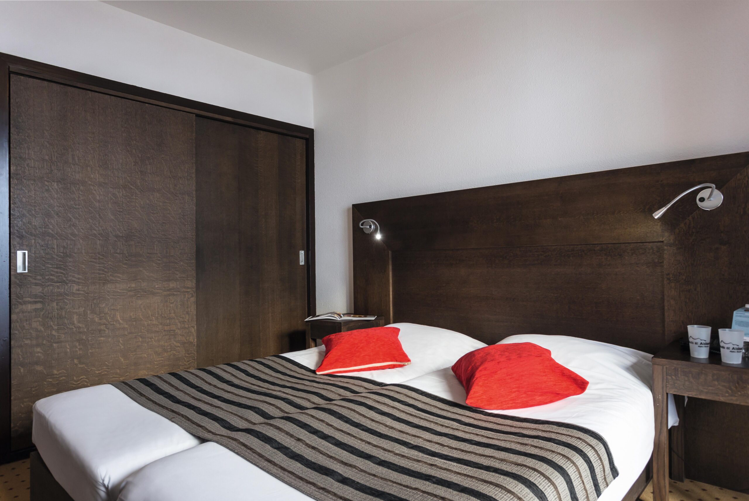 Hotelværelset, perfekt til et skiferie-retreat, har en pænt lavet dobbeltseng med en sengegavl i mørkt træ. Sengen er prydet med et stribet tæppe og to røde puder. Blød belysning og en træskydedør fuldender den hyggelige alpine atmosfære.
