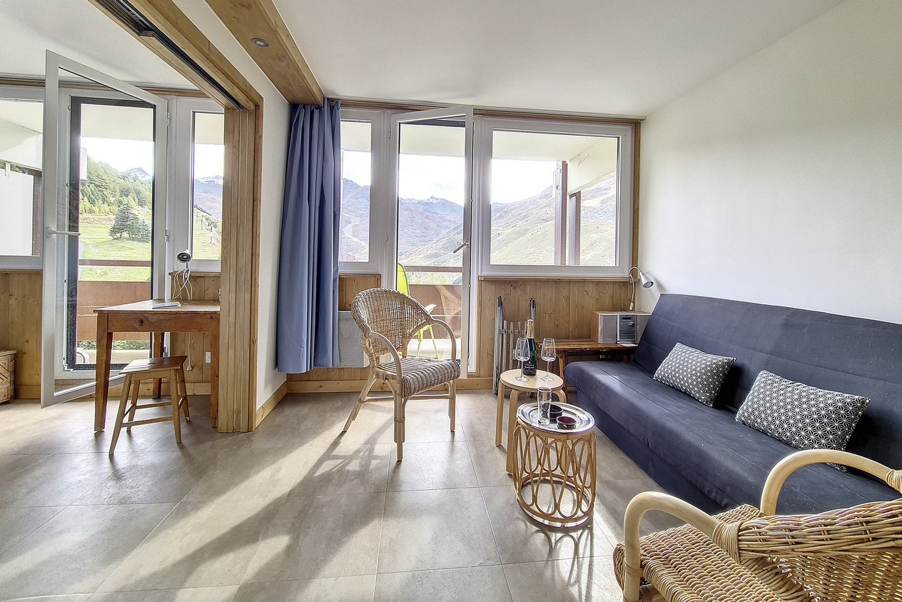 En hyggelig stue med store vinduer med udsigt over bjergene. Den har en sort futon, kurvestole, et lille træbord og et skrivebord. To glas og en flaske står på bordet, og rummet har en naturlig og indbydende stemning.