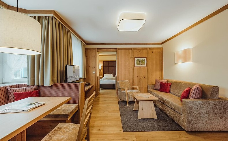 Hyggelig hotelsuite med træinteriør med opholdsområde med brun sofa, røde puder og lænestole. Et lille spisebord med stole er til venstre, og en seng er synlig gennem den åbne døråbning. Varm belysning skaber en indbydende atmosfære.