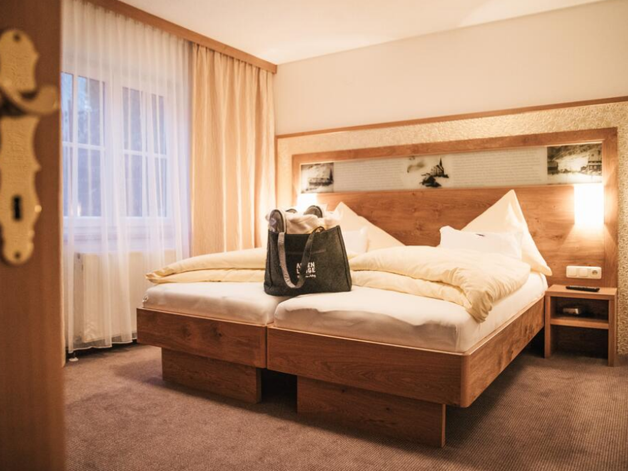 Et hyggeligt hotelværelse med en pænt lavet dobbeltseng med lyst beige sengetøj. Et stort vindue med gennemsigtige gardiner giver naturligt lys. En lille mulepose hviler på sengen, og der er kunstværker over sengegavlen. Rummet har en varm og indbydende atmosfære.