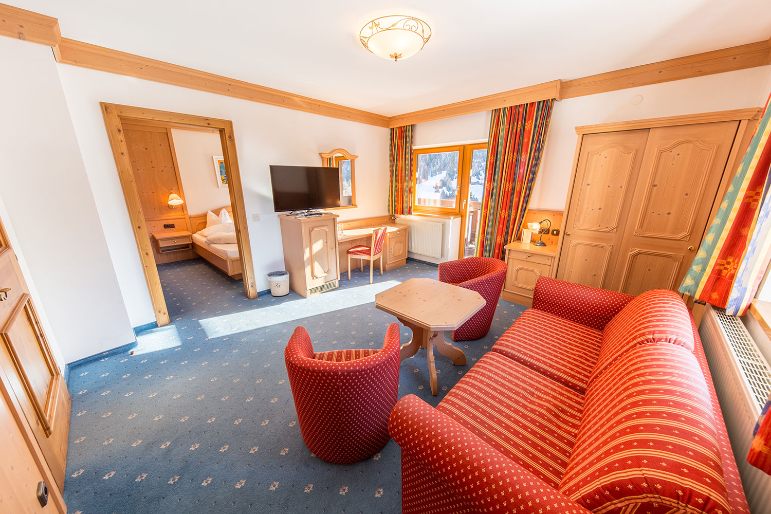 Et hyggeligt hotelværelse med en rødstribet sofa, to røde lænestole og et sofabord i træ. Et fladskærms-tv er monteret på væggen. Lyse gardiner indrammer vinduet, og en døråbning fører til et soveværelse med en seng. Indretningen er varm og rustik.