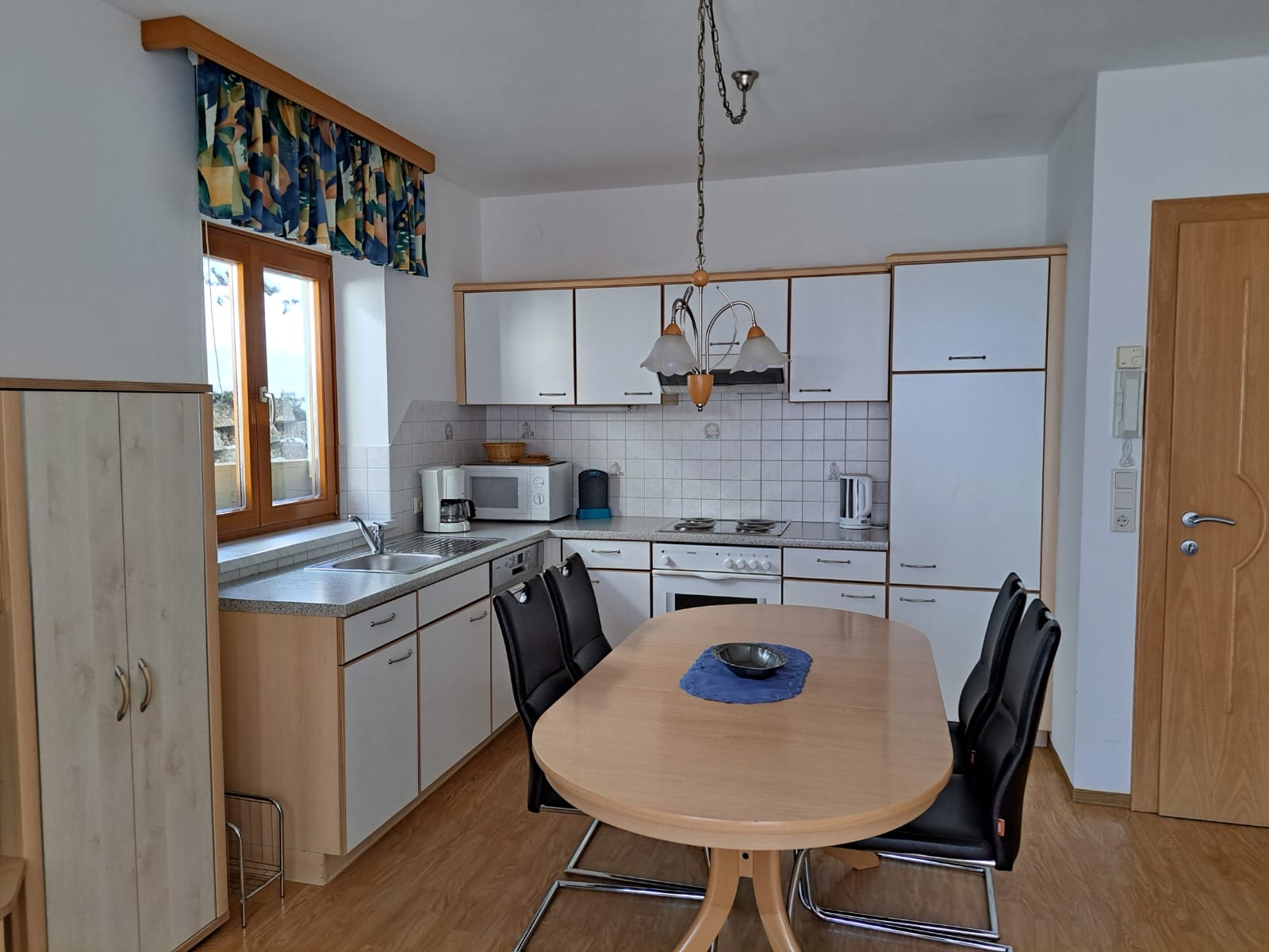 Et køkken med hvide skabe, et spisebord i træ omgivet af fire sorte stole og et vindue med et farverigt gardin. Der er apparater som mikroovn, kedel og komfur til stede. En skål med et klæde står på bordet. Gulvet er træpaneler.