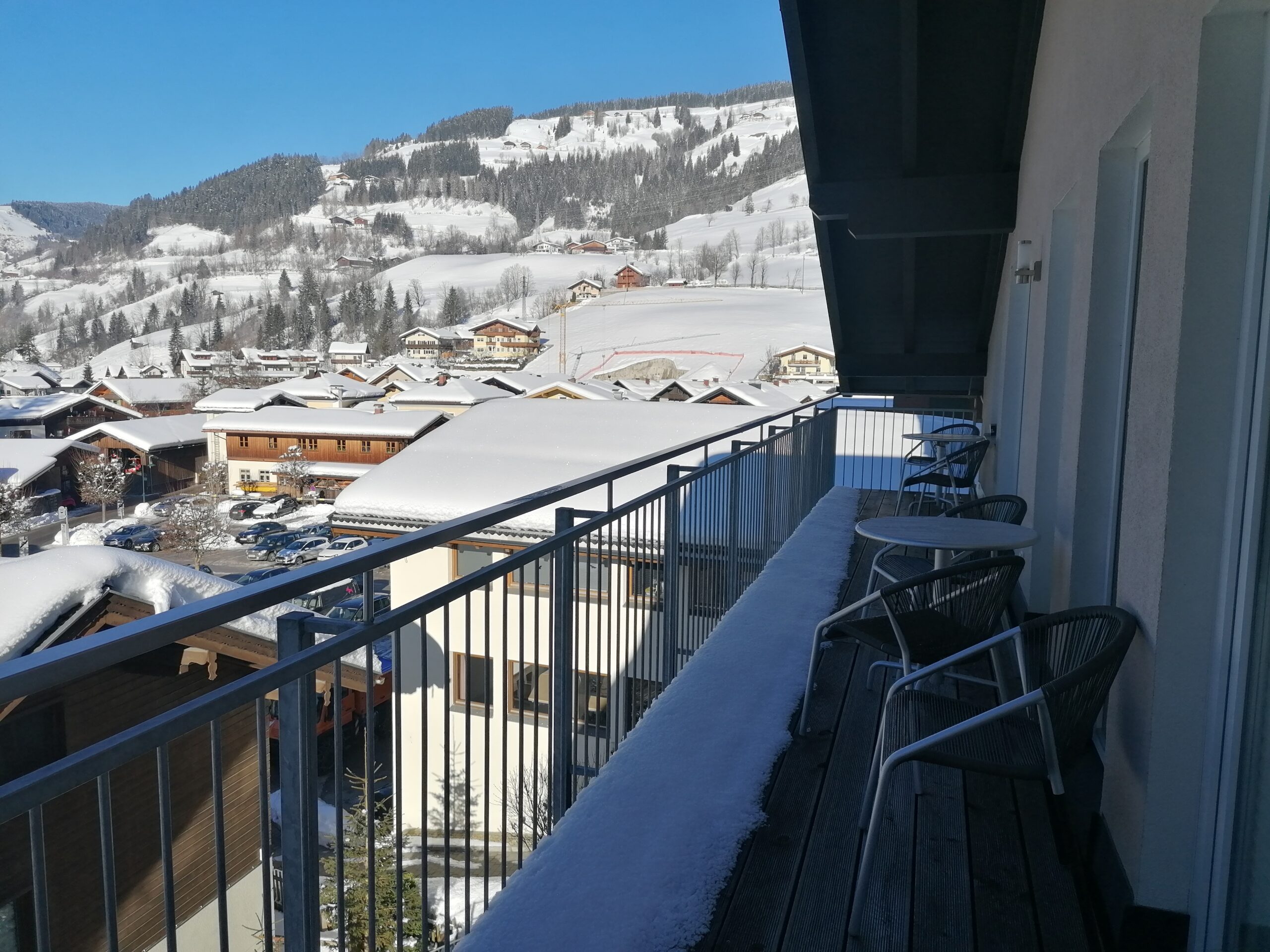 Udsigt fra en balkon med snedækkede rækværk, med udsigt over en landsby med snefyldte tage. Baggrunden har skovklædte bakker og klar blå himmel, hvilket skaber et fredfyldt vinterlandskab.
