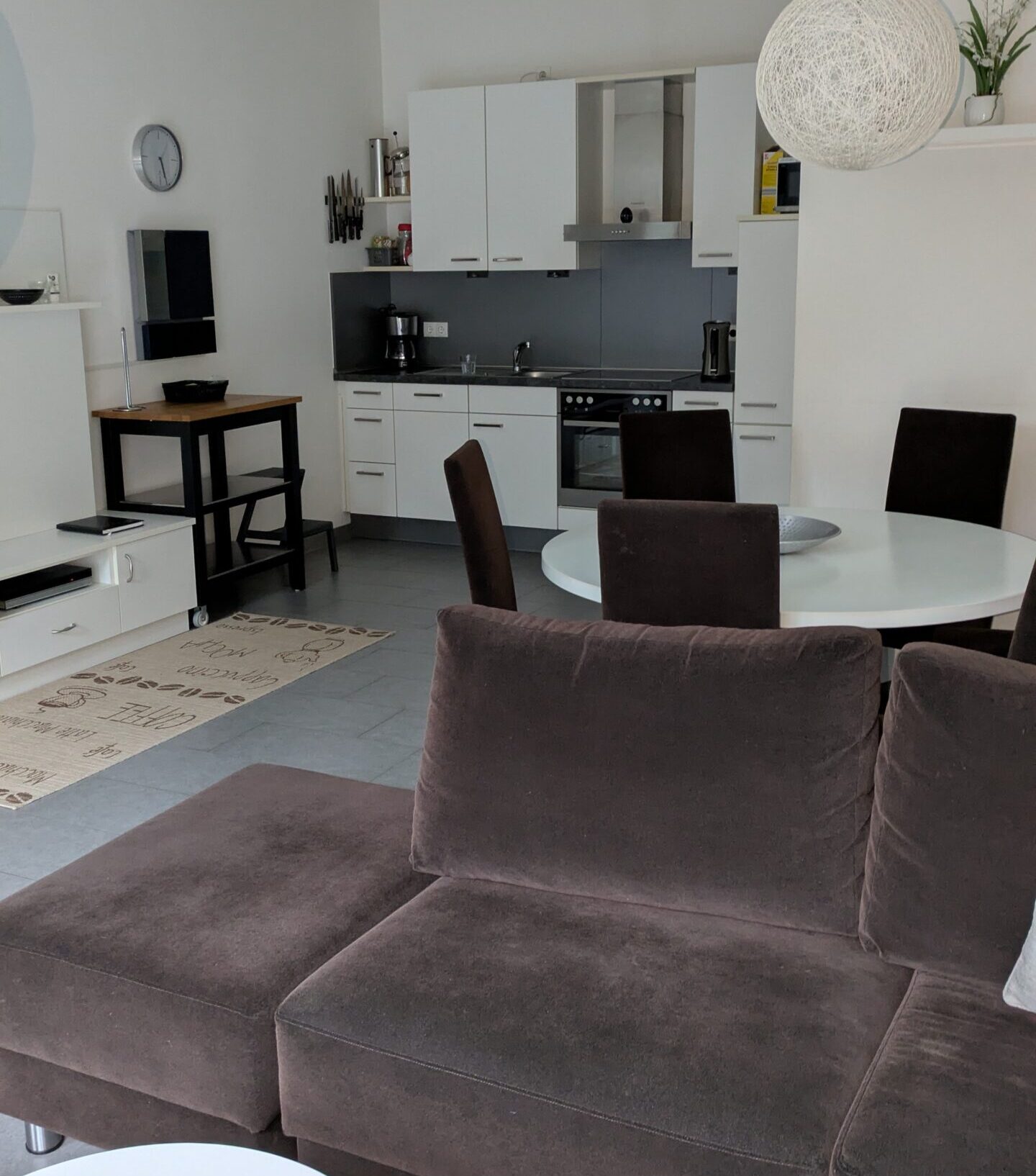 Moderne åbent stue og køkken med brun sofa, hvidt rundt spisebord, mørke stole, hvide skabe og en pendel. Fjernbetjening på et lille bord og et vægur er også synlige.