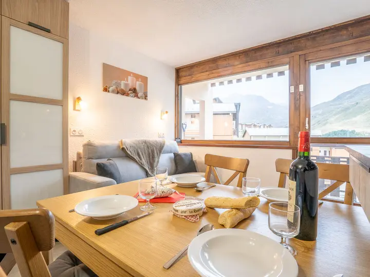 En hyggelig spiseplads har et træbord, der er dækket til et måltid med tallerkener, bestik, vin og baguetter. En grå sofa sidder i baggrunden nær store vinduer med udsigt over bjergene. Varm belysning og vægkunst med stearinlys-tema skaber en indbydende atmosfære.