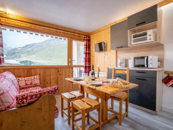 Hyggelig bjerghytte interiør med trævægge. Et spisebord er dækket med en flaske vin og brød. En sofa med rødmønstrede puder er i nærheden. Et stort vindue giver udsigt til grønne bakker. Køkkenmaskiner står på tælleren.