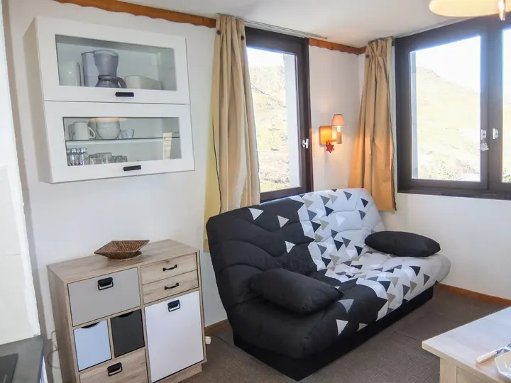 Hyggelig stue med mønstret sort/hvid sofa, træskab og væghængte hylder. Store vinduer med gardiner giver udsigt over bjergene. Væglampe giver varm belysning.