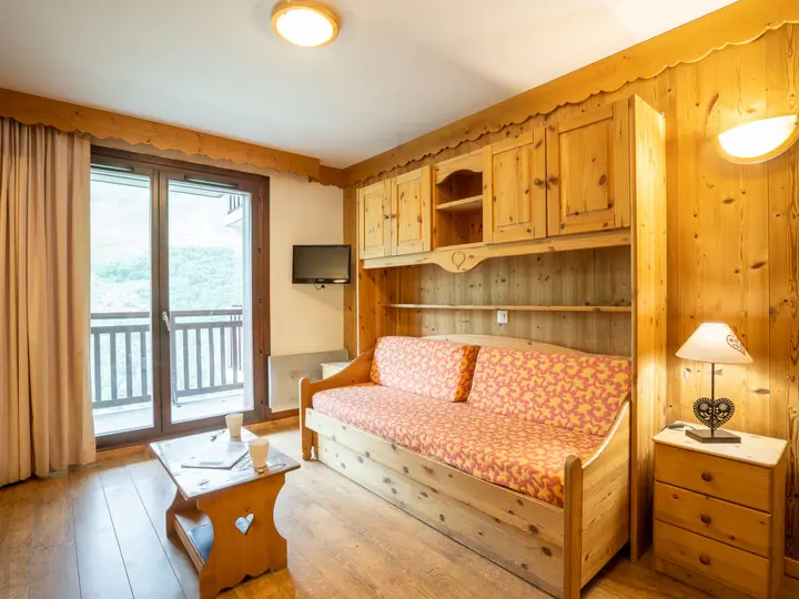 Hyggelig stue med vægge med træpaneler, en træsofa med mønstrede hynder, et lille sofabord og et natbord med en lampe. Der er et fladskærms-tv og en glasskydedør, der fører til en balkon med en naturskøn udsigt.