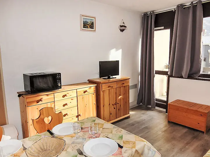 Et hyggeligt værelse med træmøbler, herunder kommode, lille bord og bænk. En mikrobølgeovn og tv sidder på kommoden. Der er et vindue med grå gardiner, et lille maleri og et bord dækket med tallerkener og glas på en farverig dug.