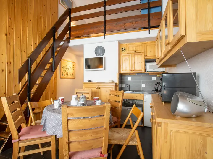 Hyggelig spiseplads i træpanelerum med bord og stole. En hems med rækværk er over. Tekøkkenet har træskabe, mikrobølgeovn og lille køleskab. Et vægur og TV er monteret på væggen. En radio sidder på en skænk.