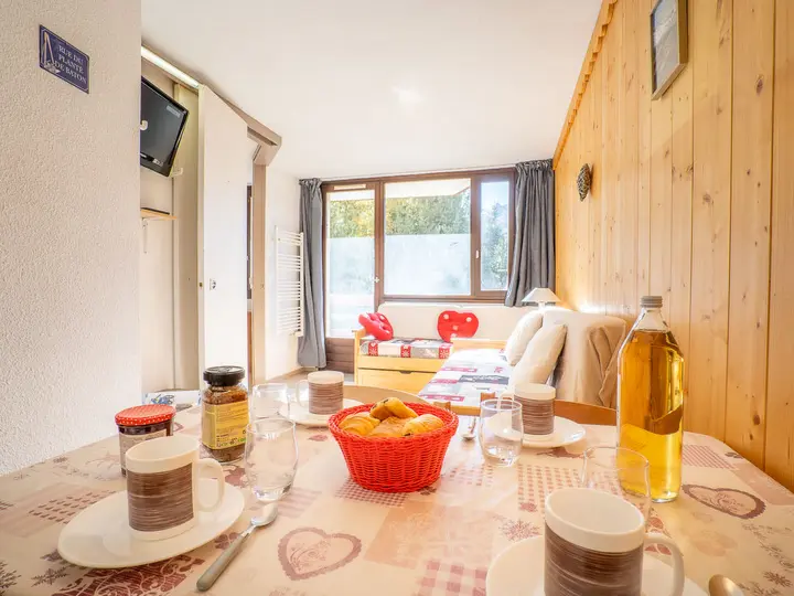 Hyggeligt værelse med trævægge og et stort vindue, der viser grønne omgivelser udenfor. Et bord er dækket med kopper, vandglas, en kurv med kager, marmelade og en flaske juice. En sofa og en seng med hjerteformede puder er i baggrunden.