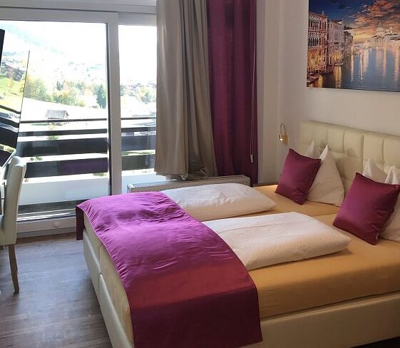 Moderne hotelværelse med dobbeltseng, magenta-accenter, hvide møbler, et vægmonteret tv, skrivebord og stol ved et stort vindue og balkon med udsigt over bjerge og grønne områder.