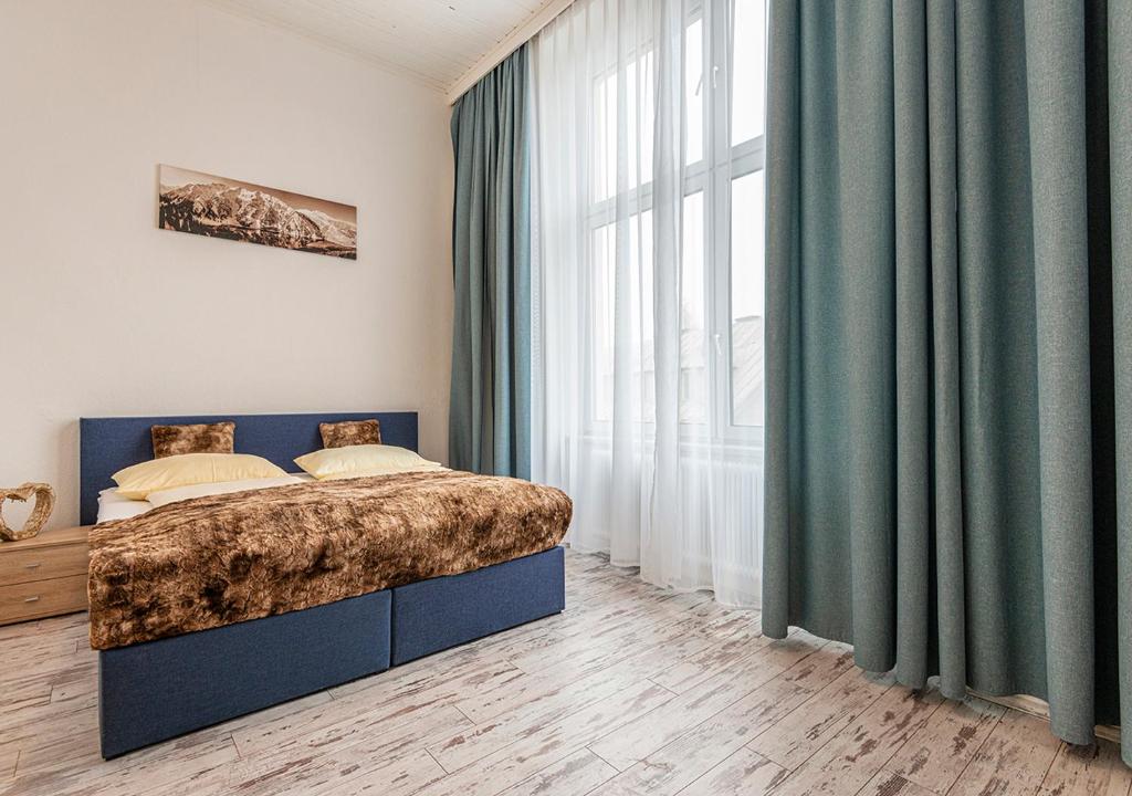 Et moderne soveværelse med en stor seng med brunt pelssengetøj og gule puder. Rummet har trægulv, blå gardiner på et højt vindue, et lille natbord og et bjerglandskabsbillede på en lys væg.