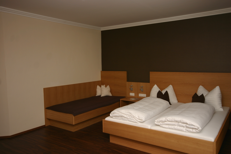 Et minimalistisk hotelværelse med en dobbeltseng og en enkeltseng, begge med trærammer, hvidt sengetøj og brune accentpuder, sat mod neutrale vægge og mørkt trægulv.