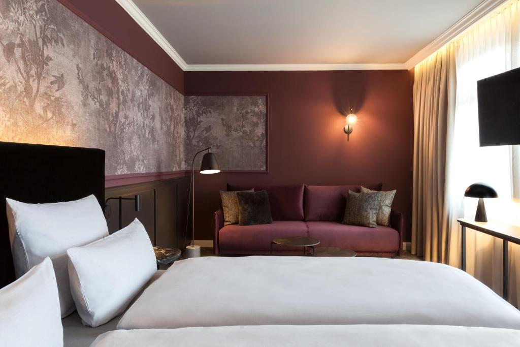 Hotelværelse med to enkeltsenge, hvidt sengetøj, bordeaux vægge og vægkunst med skovtema. En bordeaux sofa står op ad væggen med plyspuder. En gulvlampe, væglampe og bordlampe giver belysning. Et vindue lukker naturligt lys ind.