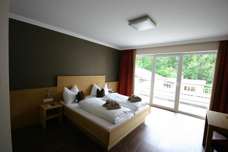 Et moderne hotelværelse med en dobbeltseng, hvidt sengetøj, brune accentpuder, natborde, et skrivebord og store vinduer med røde gardiner med udsigt over det grønne udenfor.