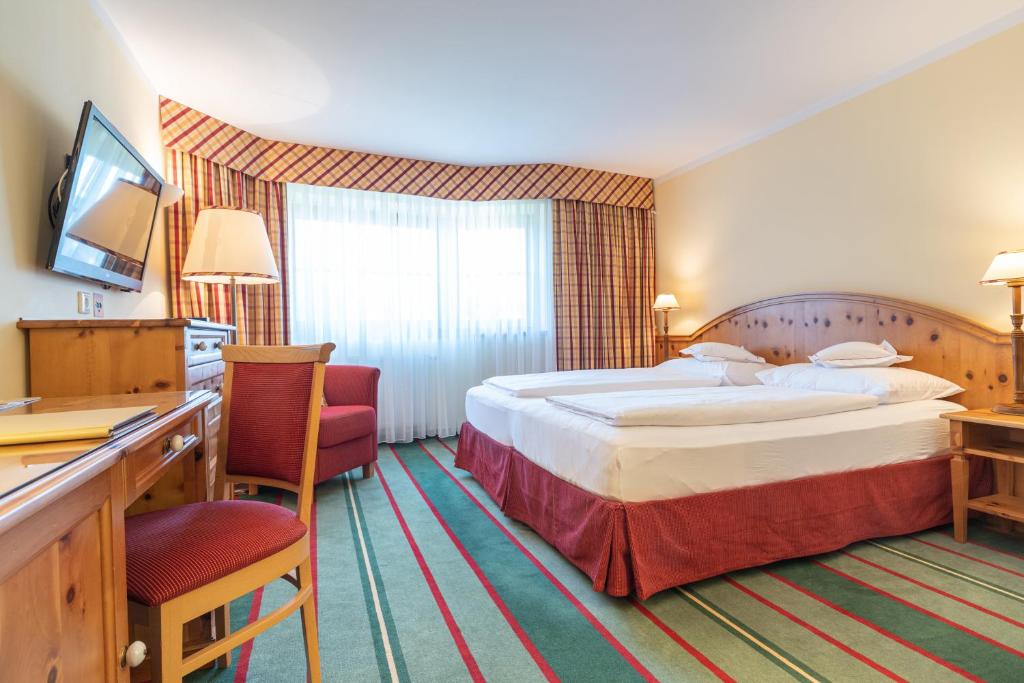Hotelværelse med king-size seng og rød- og grønstribet tæppe. Træmøbler inkluderer et skrivebord med en stol og et sidebord med en lampe. Værelset har væghængt tv og stort vindue med stribede gardiner. Varm belysning skaber en hyggelig atmosfære.
