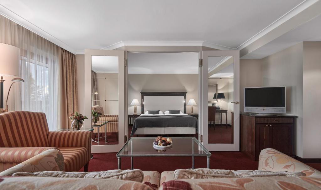 Et rummeligt hotelværelse med en king-size seng og to spejle på hver side. Der er en stribet lænestol og en blomstret sofa i forgrunden. Et glas sofabord og et tv på et træskab fuldender de hyggelige rammer.