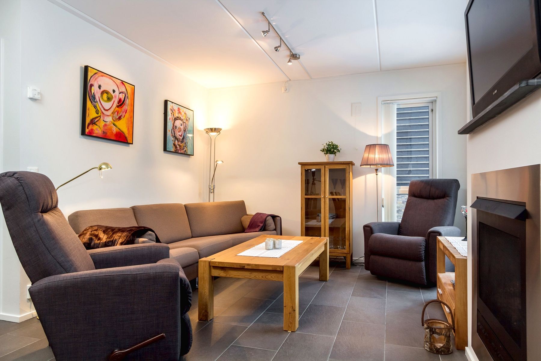 En hyggelig stue med en brun sofa, to mørke lænestole, et sofabord i træ, et væghængt tv, dyrekunst og et skab med glasfront, varmt oplyst af gulv- og bordlamper.