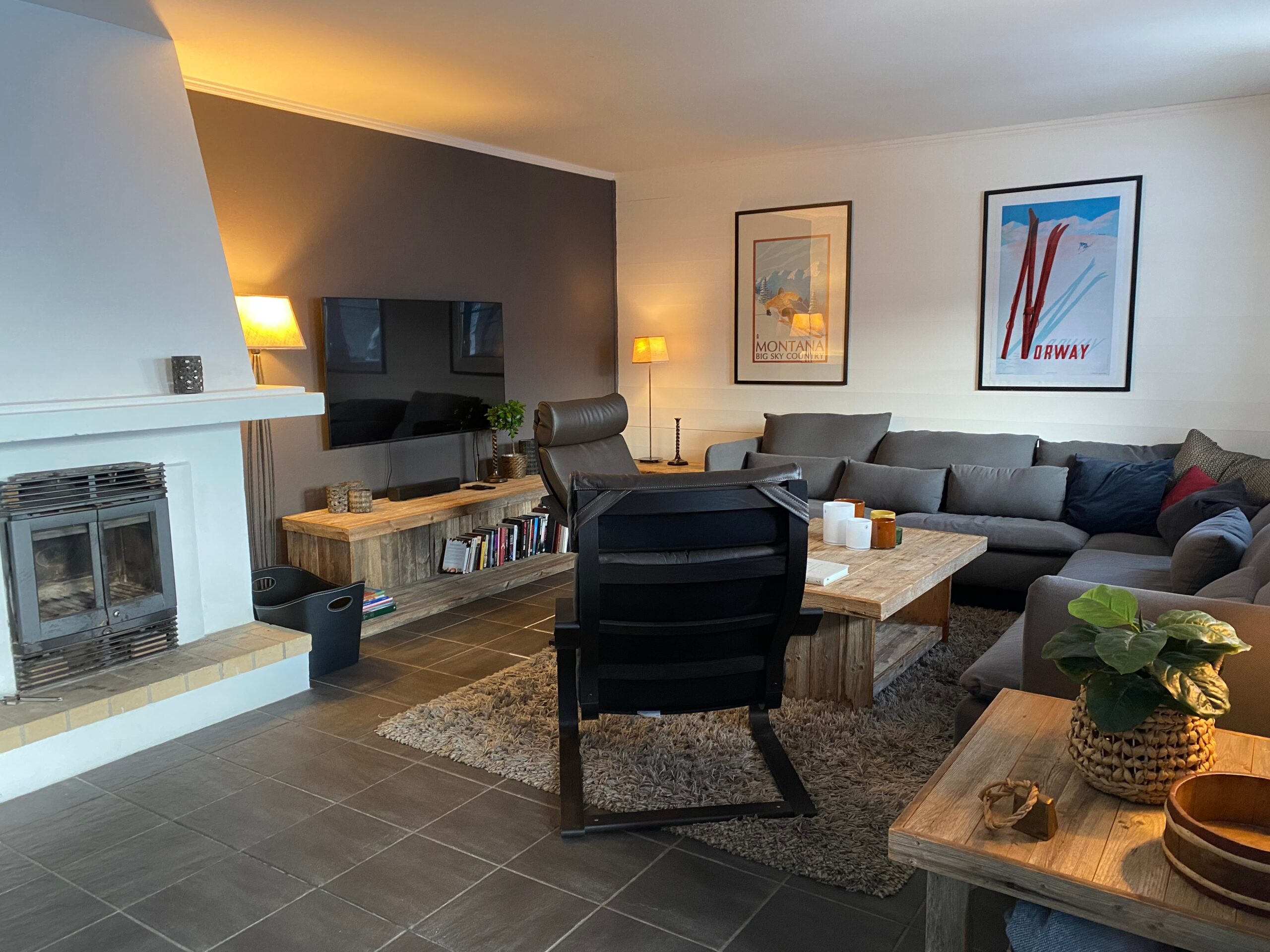 Hyggelig stue med en grå hjørnesofa, sofaborde og sideborde i træ, en sort lænestol, pejs, TV og kunst på væggene. Varm belysning og stueplanter skaber en behagelig atmosfære.