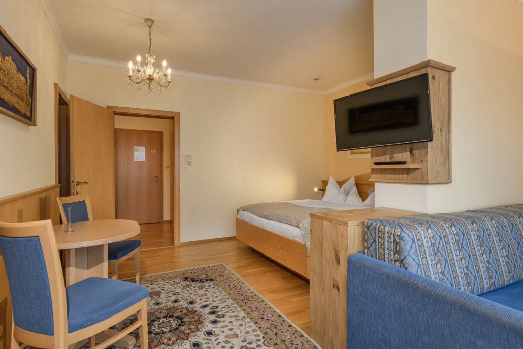 Hotelværelse med seng, sofa og lille spisebord med to stole. Et tæppe er på trægulvet, og en lysekrone hænger fra loftet. Et fladskærms-tv er monteret på væggen. Væggene er lyse med et billede og træaccenter.