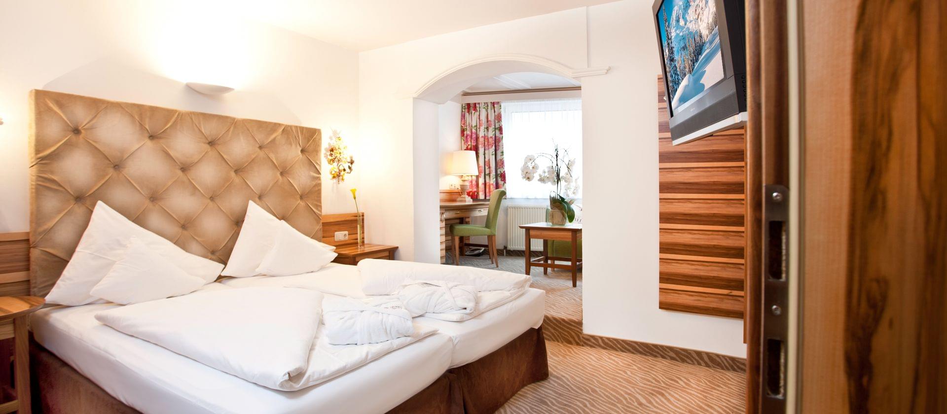 Et hyggeligt hotelværelse med en stor seng, hvidt sengetøj, to morgenkåber, en tuftet sengegavl, træaccenter, et skrivebord med en lampe, blomstergardiner og naturligt lys, der strømmer gennem et vindue.