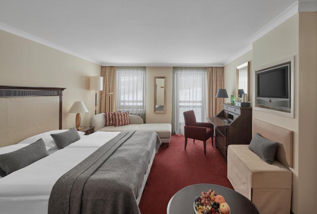 Hotelværelse med stor seng, gråt sengetøj og pyntepuder. Der er en chaiselong, lænestol, skrivebord og tv. Værelset har rød løber, beige vægge, store vinduer med gardiner og en frugtskål på et lille bord.