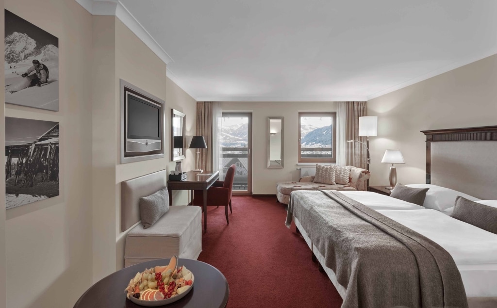 Hotelværelse med dobbeltseng, rød løber og beige vægge. Et lille bord med frugt, et fladskærms-tv og to vinduer tilbyder udsigt over sneklædte bjerge. Hyggelige siddepladser nær vinduerne, og sort-hvide billeder på væggene forbedrer indretningen.
