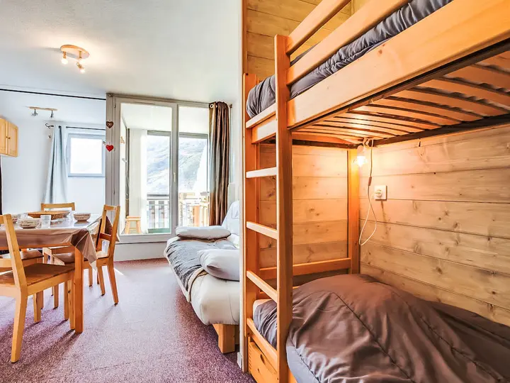 Hyggeligt værelse med trækøjesenge til højre, sofa og spiseplads til venstre. Solbelyst balkon med udsigt over bjergene synlig gennem en glasdør og vindue i baggrunden. Varme farvetoner skaber en indbydende atmosfære.