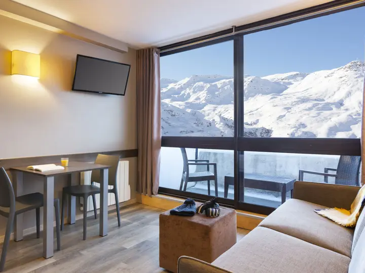 Moderne hotelværelse med et stort vindue med en malerisk udsigt over sneklædte bjerge. Indeni er der en sofa, et lille spisebord med stole, et vægmonteret tv og et par handsker på en polstret fodskammel.