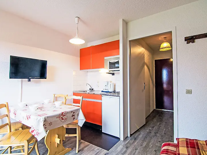 Et lille køkkenområde med lys orange skabe, et væghængt tv og et spisebord af træ dækket med en mønstret dug. Rummet har træstole og en gang, der fører til en lukket dør. En pendel hænger fra loftet.