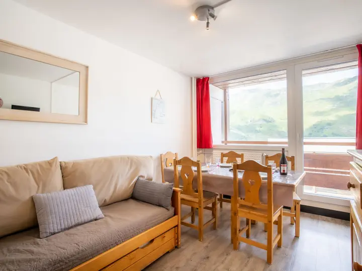 En hyggelig spiseplads med et træbord til seks, flankeret af røde gardiner. En beige sofa med grå hynder er til venstre, og store vinduer viser en naturskøn udsigt over bjergene i baggrunden. Et spejl hænger på væggen over sofaen.