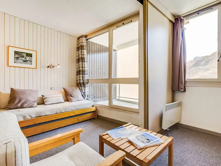 Hyggelig stue med minimalistisk design, med en beige sofa, trælænestol og et lille sofabord i træ. Store vinduer med gardiner tilbyder udsigt over bjergene. Et indrammet billede hænger på den stribede væg. Neutrale toner skaber en varm atmosfære.