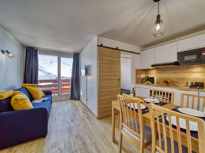 Moderne lejlighedsinteriør med en hyggelig opholdsstue med en blå sofa og gule puder. Et spisebord til fire er tæt på det kompakte køkken. Store vinduer afslører et snedækket bjerglandskab udenfor. Varm belysning og træaccenter skaber en indbydende stemning.