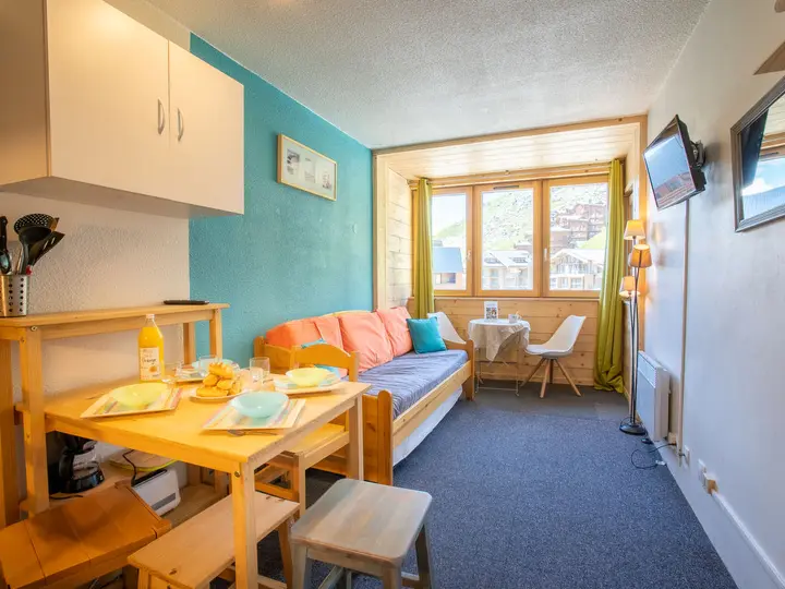 Et hyggeligt opholds- og spiseområde med et lille træbord til tre, en sofa med orange hynder, et vindue med grønne gardiner, et væghængt tv og et stærkt naturligt lys, der fylder rummet.
