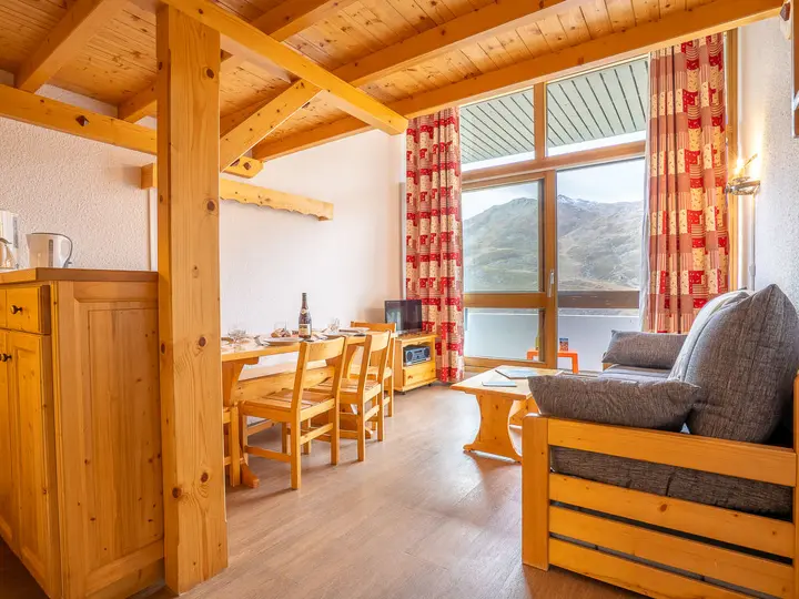 Hyggeligt hytteinteriør med træmøbler, herunder sofa, bord og stole. Stort vindue med bjergudsigt og rødmønstrede gardiner. Varm belysning, en kedel på disken og en flaske på bordet forstærker den indbydende atmosfære.