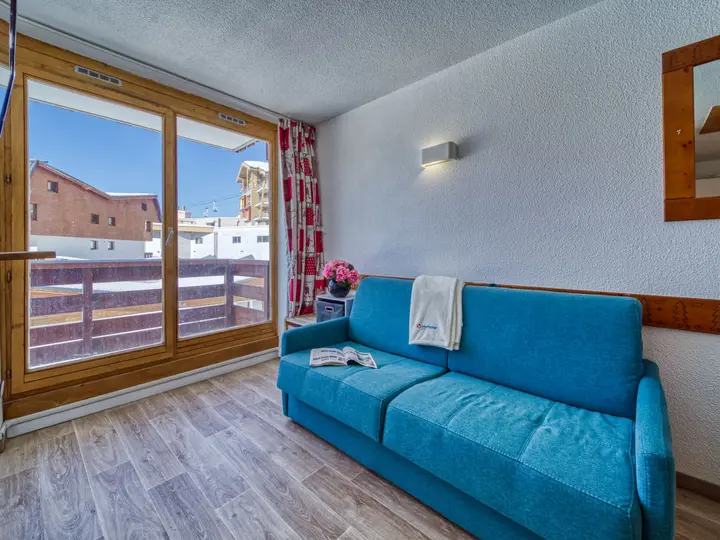 Et hyggeligt værelse med en blå sofa mod en hvid væg, dekoreret med et spejl og en væglampe. Et lille bord med blomster og en bog i nærheden. Stort vindue afslører snedækkede bygninger og en klar blå himmel udenfor. Røde og hvide gardiner hænger ved vinduet.